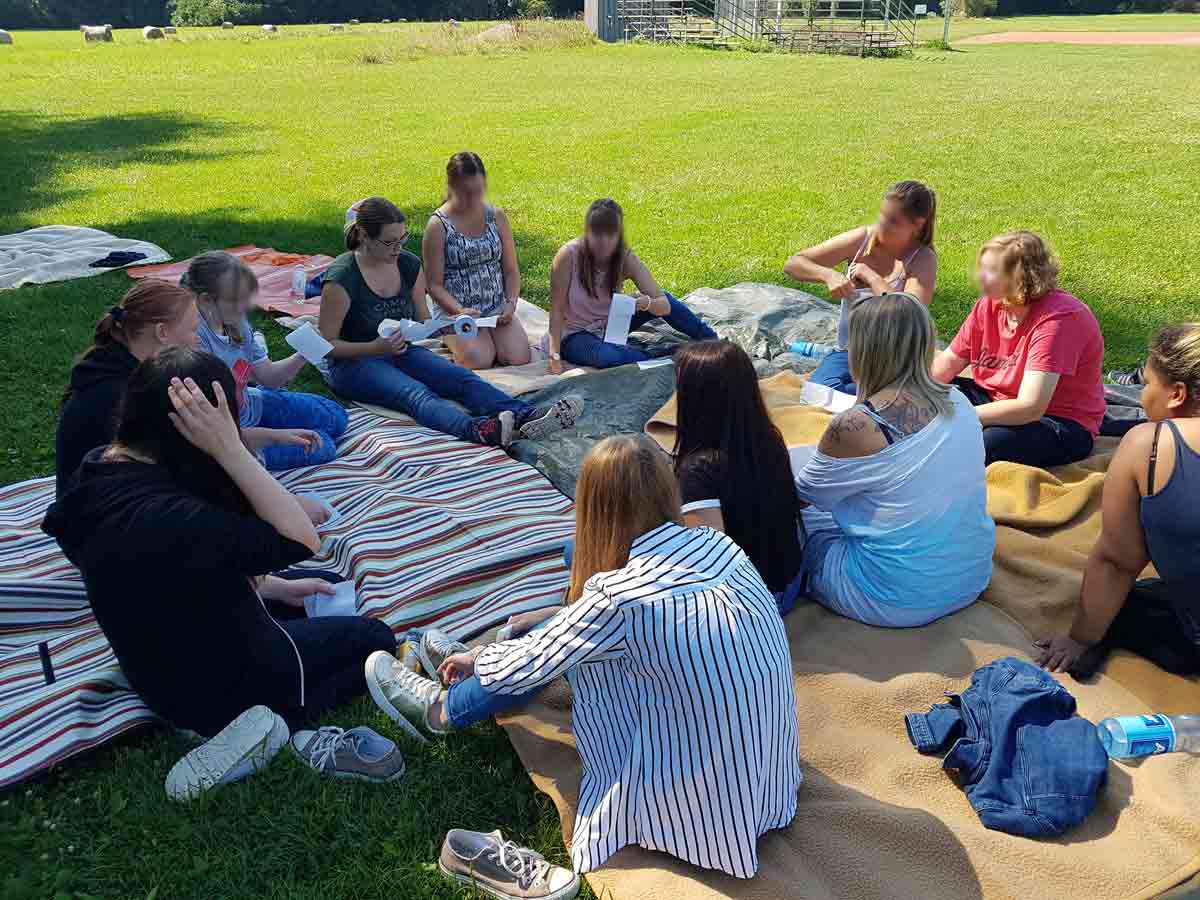 Eine Gruppe junger Erwachsener sitzt im Kreis auf einer Wiese während eines Workshops zur Teamentwicklung und tauscht sich über ihre Aufgaben aus.