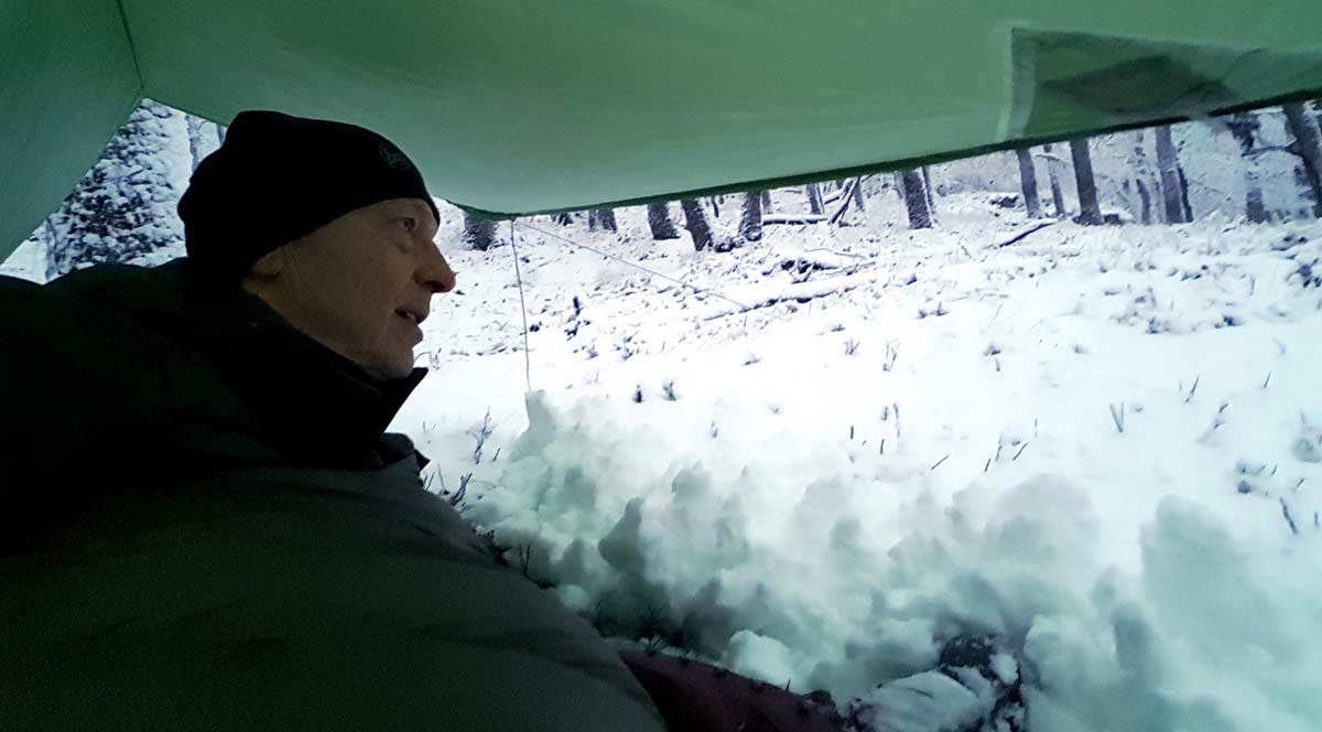 Ein Mann (Uwe Kreß) liegt im Winterwald in einem Schlafsack direkt auf dem Boden und ist von einer dicken Schicht Neuschnee bedeckt. Das Bild symbolisiert Rückzug, Kälteanpassung und die psychologische Erfahrung der Raunächte für Hochsensible.