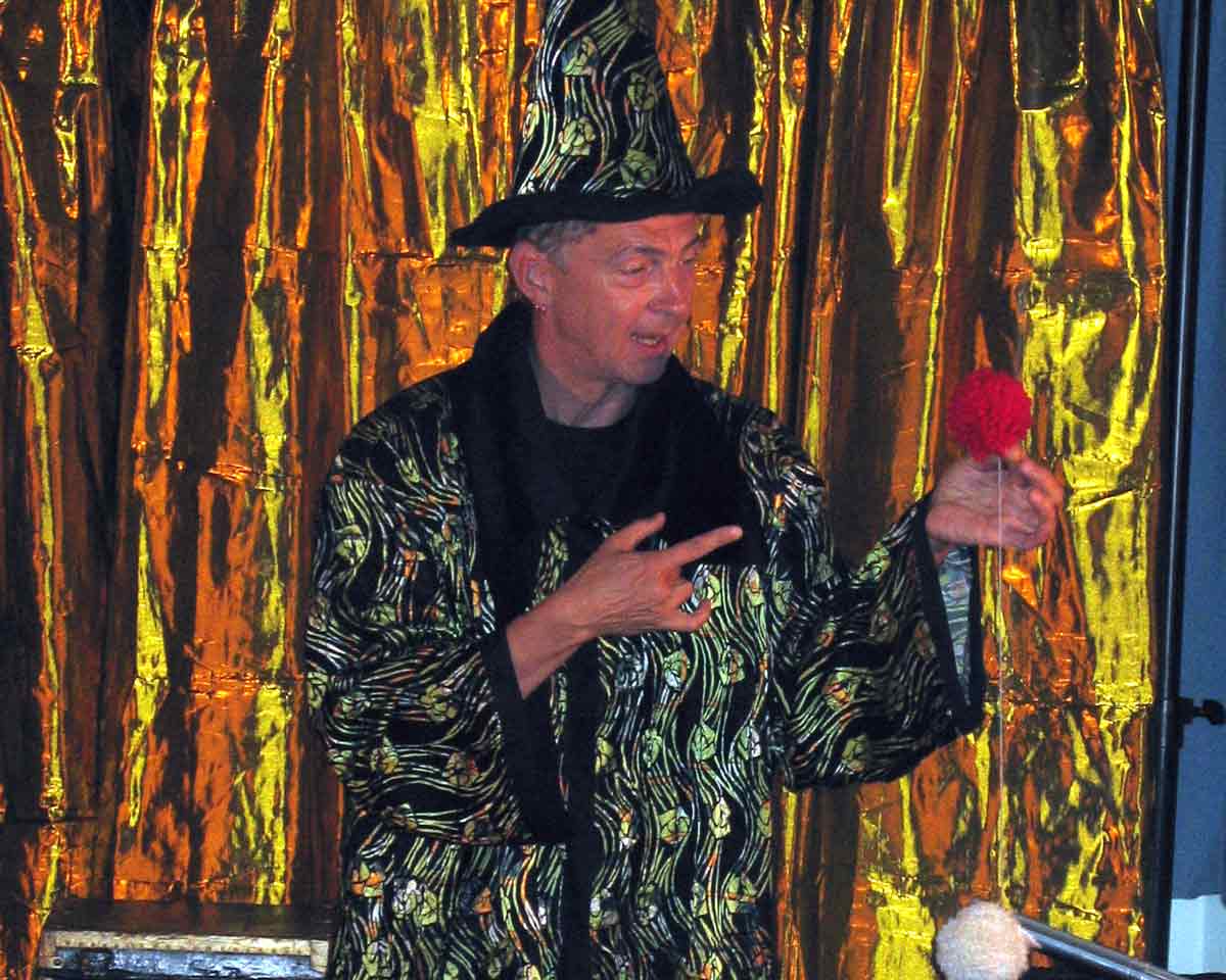 uwe-kress-buehnenmagier-aufmerksamkeit Götz Uwe Kreß als Bühnenmagier mit Hut und Robe, eine rote Blume in der erhobenen Hand, vor goldenem Vorhang