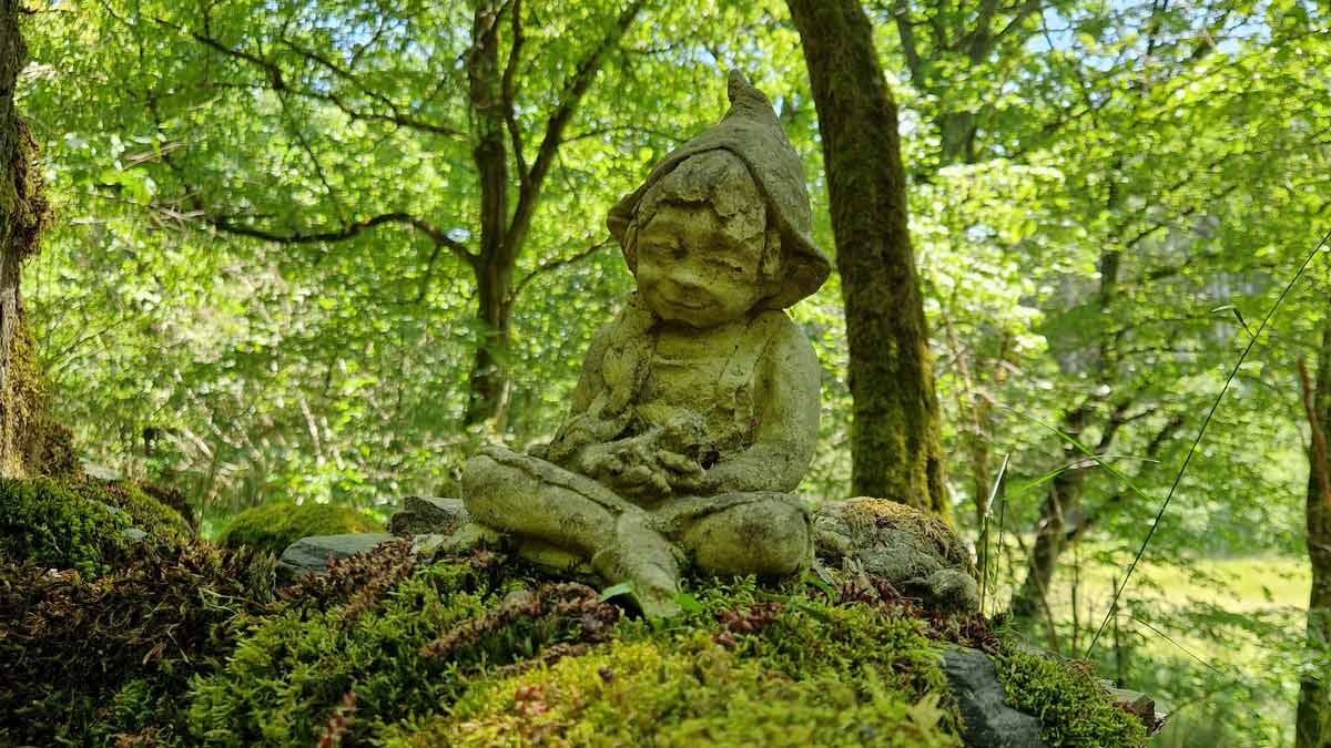 Steinerner Wichtel im Wald Steinfigur eines sitzenden Wichtels mit Zipfelmütze auf moosbewachsenem Stein im lichten Wald
